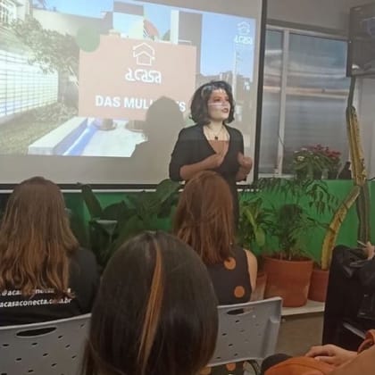 Raisa Arruda apresentando diante de uma plateia sentada, com slide projetado ao fundo.