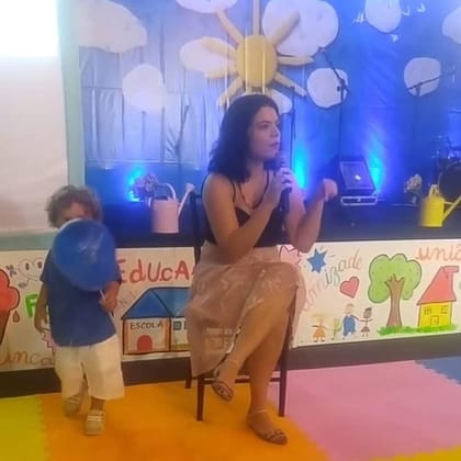 Raisa Arruda falando ao microfone em palestra dentro de uma escola, com uma criança em primeiro plano.