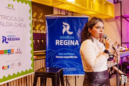 Raisa Arruda ao microfone durante palestra em evento, com banners dos patrocinadores ao fundo.